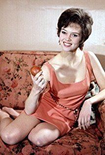 Gabrielle Drake with a smiling face while sitting on an orange couch, with short hair, wearing a sexy sleeveless orange dress and holding a red apple.