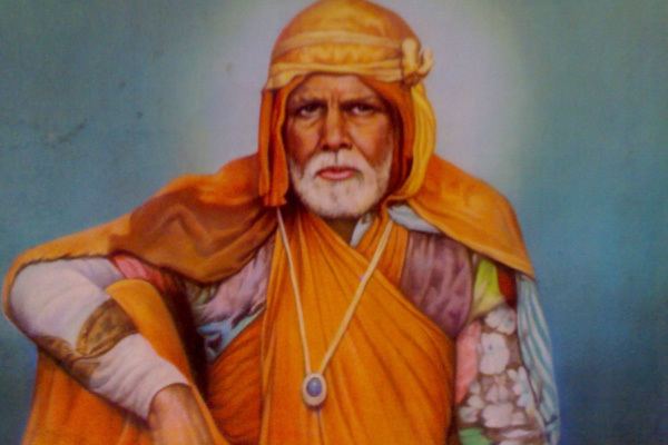 Gadge Maharaj with a serious face while his arm is on his leg and with a grey beard while wearing a long gold necklace and a turban, traditional Indian clothing