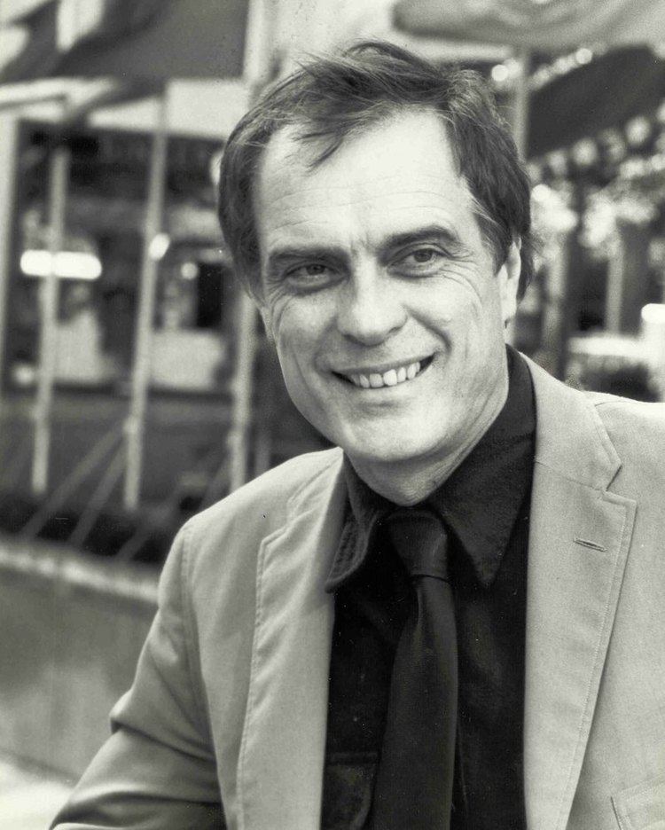 Gardner McKay smiling while wearing a coat, black long sleeves, and black necktie
