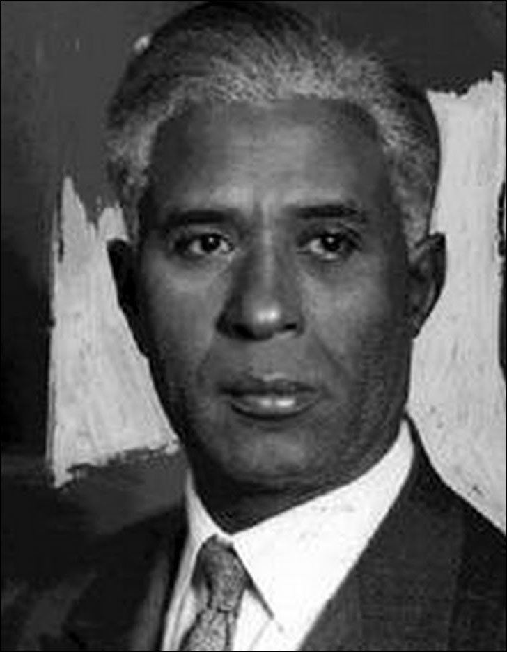 Garrett Augustus Morgan, Sr. with a serious face, wearing a black coat over white long sleeves and a necktie.