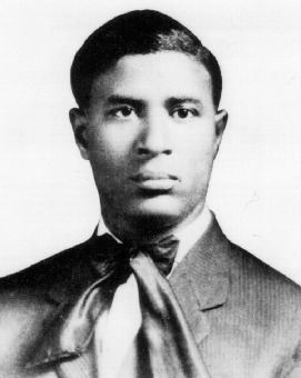 Garrett Augustus Morgan, Sr. with a serious face, wearing a coat over white long sleeves and a ribbon bowtie.