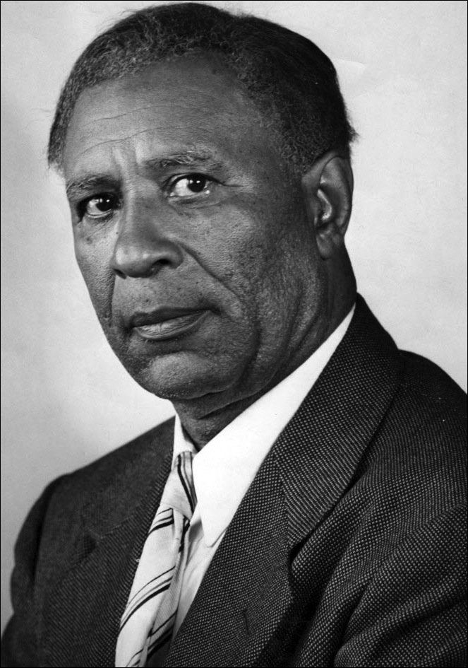 Garrett Augustus Morgan, Sr. with a serious face, wearing a black coat over white long sleeves and a striped necktie.