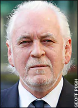 Gary Brooker with a serious face, mustache, and beard while wearing a white long sleeve under a black necktie and black coat