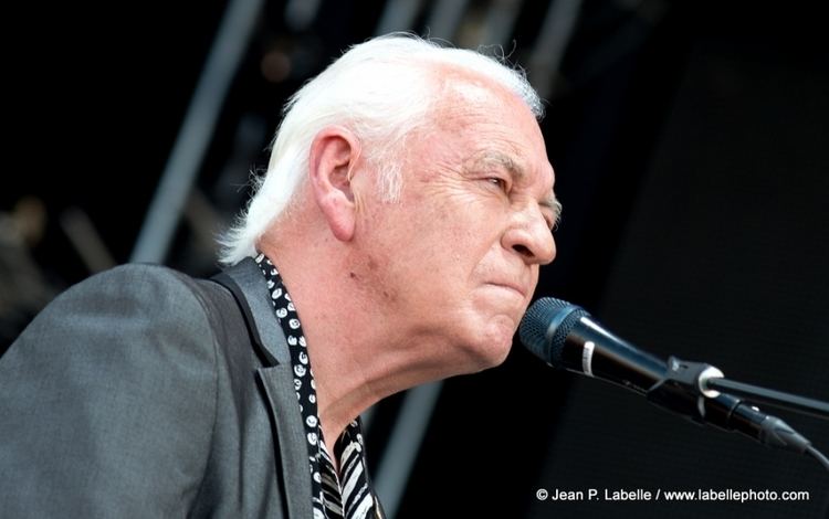 Gary Brooker singing with a serious face and white hair while wearing a black and white long sleeve under a dark gray coat