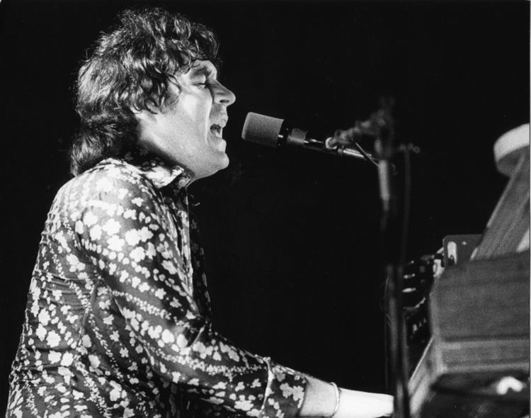 Gary Brooker singing while playing the piano with short curly hair and wearing a patterned long sleeve and bracelet