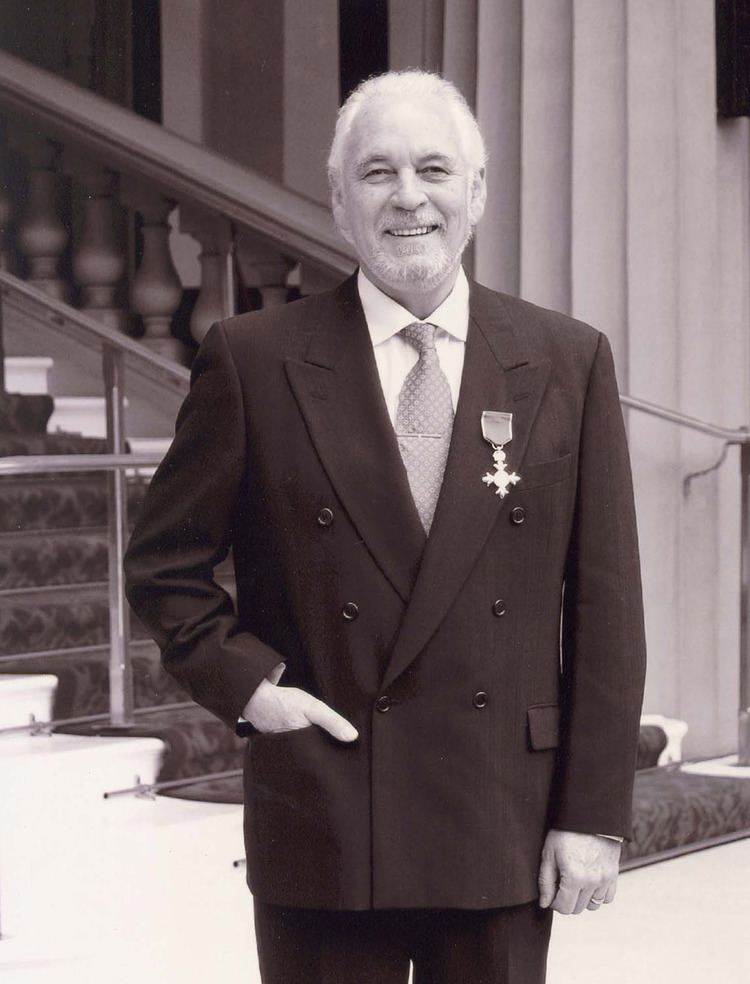 Gary Brooker smiling while hand in his pocket, with mustache and beard, and wearing brown pants and a white long sleeve under a gray necktie and brown coat with a badge