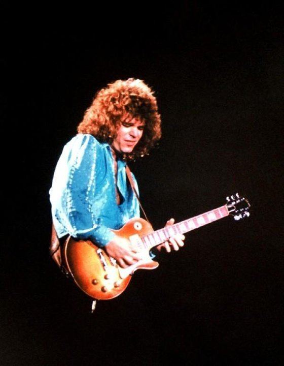 Gary Richrath smiling while playing guitar, with curly hair, and wearing a blue long sleeves shirt.