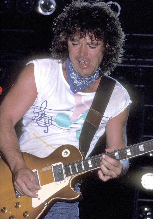 Gary Richrath playing guitar, with curly hair, with a blue handkerchief on his neck, wearing a sleeveless white shirt, and blue pants.