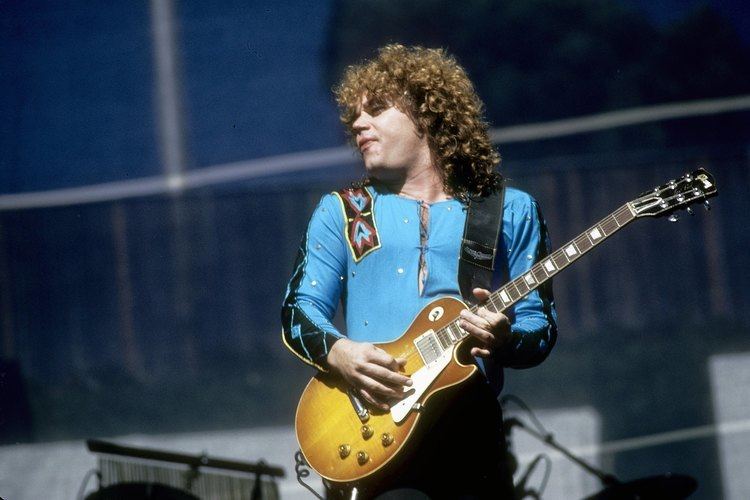 Gary Richrath with closed eyes while playing guitar, with curly hair, wearing a blue long sleeves shirt, and black pants.