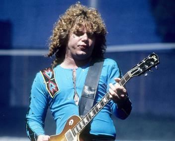 Gary Richrath with a serious face while playing guitar, with curly hair, and wearing a blue long sleeves shirt.