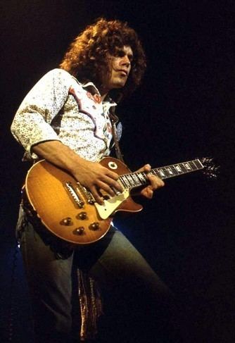 Gary Richrath with a serious face while playing guitar, with curly hair, wearing long sleeves, and black pants.