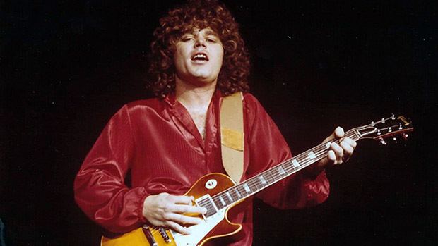 Gary Richrath sings while playing guitar, with curly hair, and wearing red long sleeves.