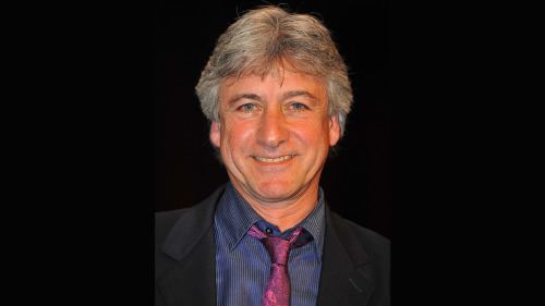 Gary Warren smiling while wearing a black coat, blue long sleeves, and violet necktie