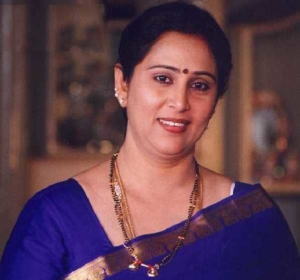 Geetha smiling with a red mark on her forehead and wearing a blue saree with beaded necklaces and earrings