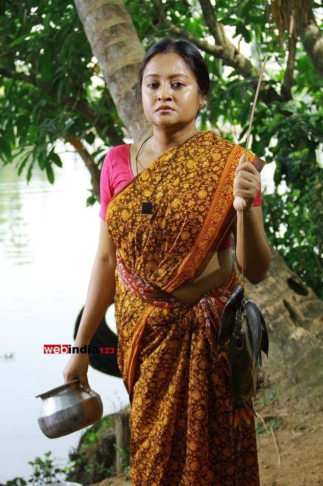 Geetha Vijayan with a serious face, wearing earrings, a necklace, and pink and orange traditional Indian clothing for women while holding a stick with fish and a cooking pot beside a river.