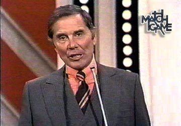 Gene Rayburn hosting the Match Game while wearing an orange long sleeve, black striped necktie, dark gray vest, and coat