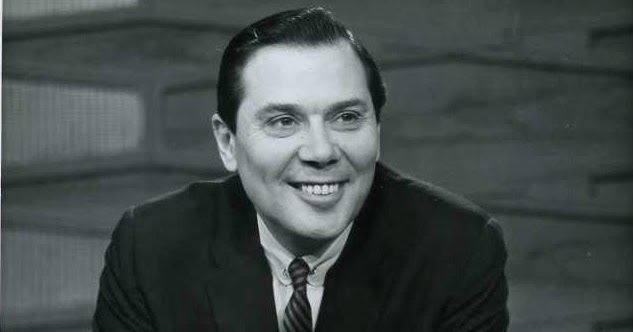 Gene Rayburn smiling while looking at something and wearing a long sleeve, necktie, and coat