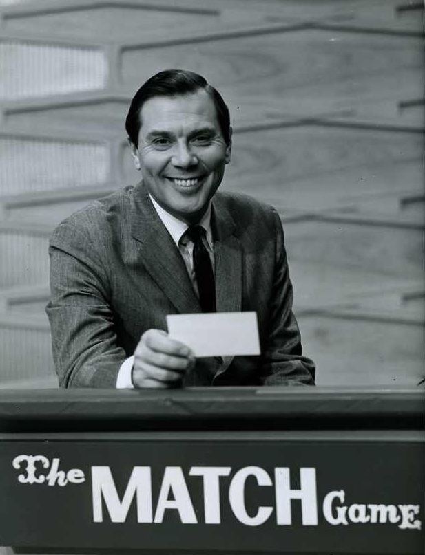Gene Rayburn smiling while hosting The Match Game and wearing a long sleeve, necktie, and coat