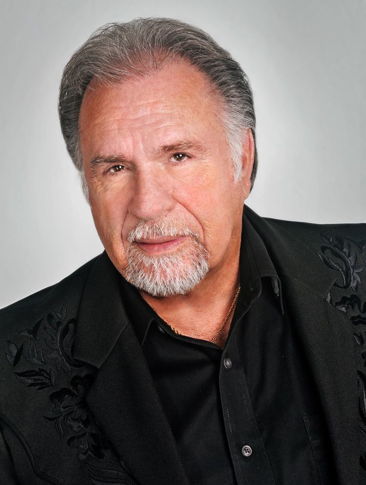 Gene Watson with a tight-lipped smile while wearing a black coat and black long sleeves
