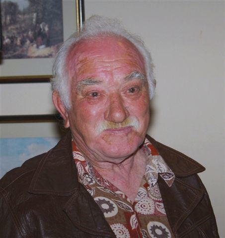 George Stafford smiling having a white hair and a white beard with a picture frame in the background and wearing a brown printed shirt under a brown leather jacket