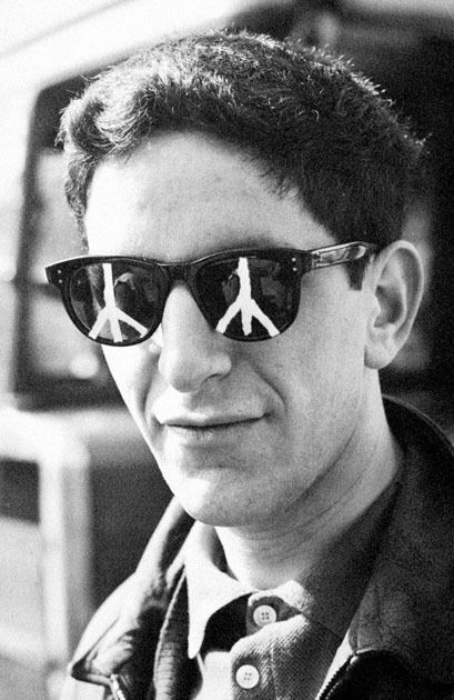 Gerald Holtom smiling wearing sunglasses with a peace sign on it and a polo shirt under a coat