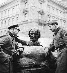 The Germans destroyed symbols of the Polish state. Two German soldiers standing near the toppled Grunwald monument in Krakow. Poland, 1940. The soldier on the left is wearing a peaked cap while the soldier on the right is wearing a beret cap and they are both wearing trench coat with a belt
