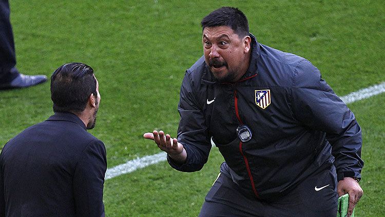 Germán Burgos with an angry face while talking to Diego Simeone inside a football ground. German with a beard and mustache, wearing a dark blue jacket and pants with the logos of Nike and Atlético de Madrid, a timer, and holding a green notebook while Diego with a beard and wearing a black coat.