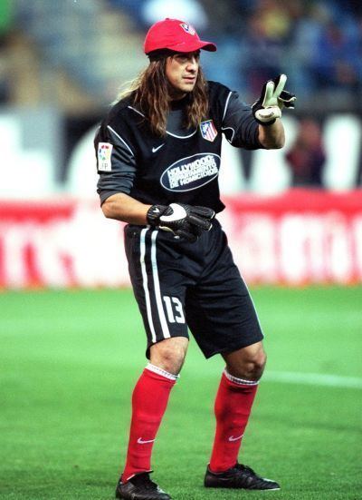 Germán Burgos doing a peace sign with a serious face inside a football ground, with long hair, wearing a red cap, a black and white football jersey and shorts with no. 13 on it, red and white socks, and black shoes.
