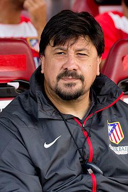 Germán Burgos sitting on the bench with a serious face, with beard and mustache, wearing a black jacket with the logos of Nike and Atlético de Madrid, and a timer.