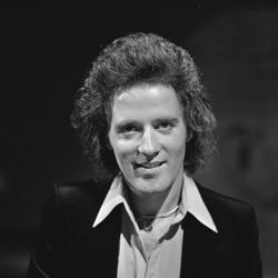 Young Gilbert O'Sullivan smiling, with curly hair, wearing a black coat over white long sleeves.