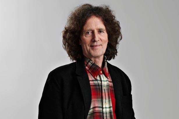 Gilbert O'Sullivan with a tight-lipped smile, curly hair, wearing a black coat over a red checkered polo shirt.