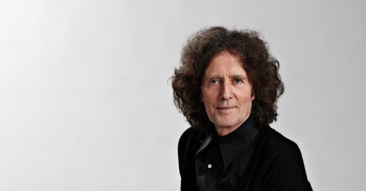 Gilbert O'Sullivan with a tight-lipped smile, curly hair, and wearing black long sleeves.
