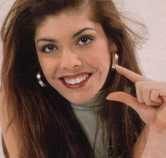 Gillette smiling while showing a hand gesture for the word small, with brown hair, and wearing hoop earrings and a green sleeveless blouse