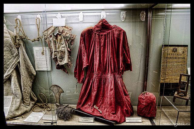 In a box of glass, from left, is a gray ragged sack, with brown knotted ropes, 2nd from left is a ragged cloth hanging on the wall, 3rd from left is a ragged red robe, 4th from left is a ragged red mask used for execution, at the right is a brown wood with old paper on it with text written with a miniature guillotine, in front is a ax with black handle and a rope wrapped in between with white papers in each item.