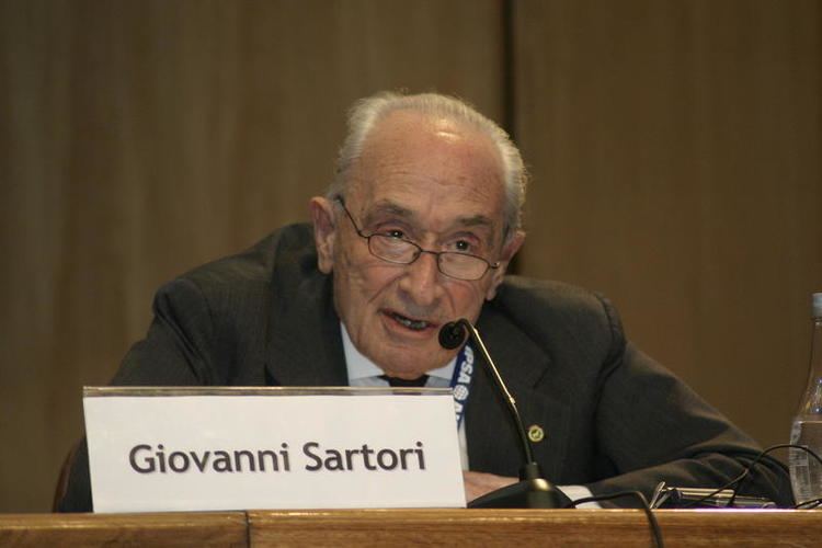 Giovanni Sartori delivering a speech while wearing a white sleeve, black coat and eyeglasses