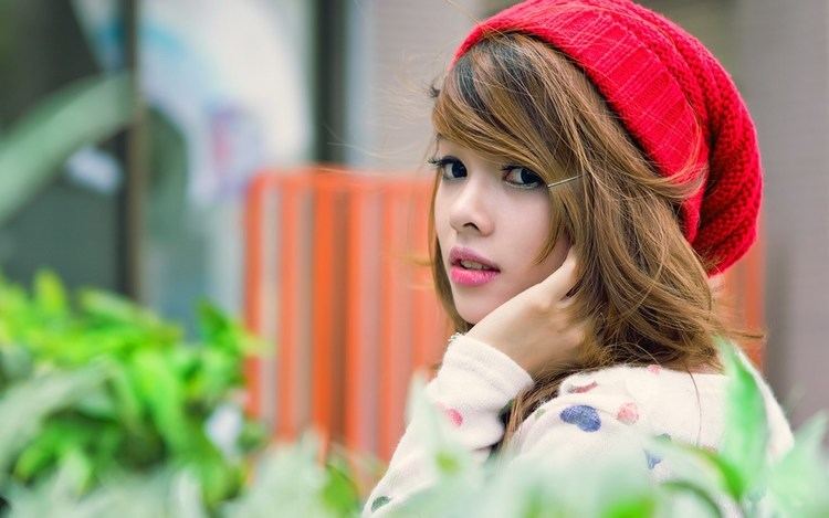 Wai Lana Butler looking from afar with her hands on her side cheeks and green leaves in her background, she has blonde hair and bangs with a pin on it, wearing a red knitted hat and a white long sleeve