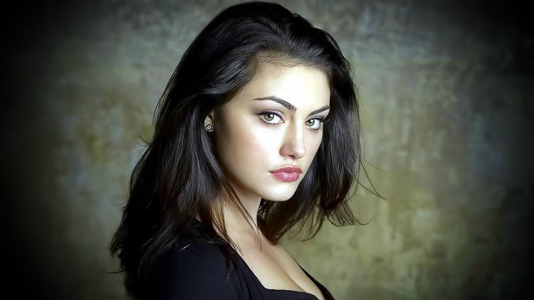 Phoebe Tonkin with a fierce look in her eyes has black hair wearing earrings and a visible cleavage black top