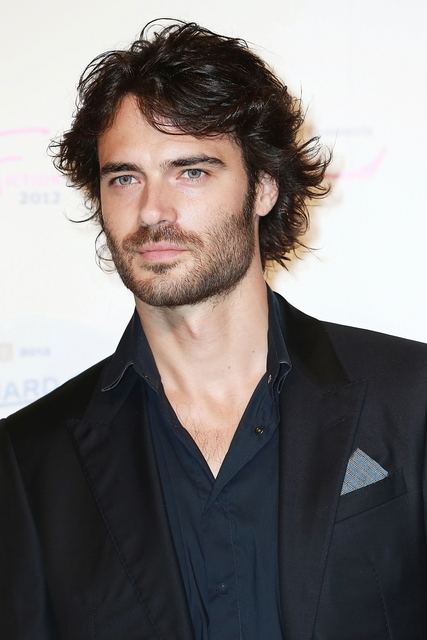 Giulio Berruti looking afar with messy hair, mustache, and beard while wearing a dark blue long sleeve under a black coat with a handkerchief in the pocket