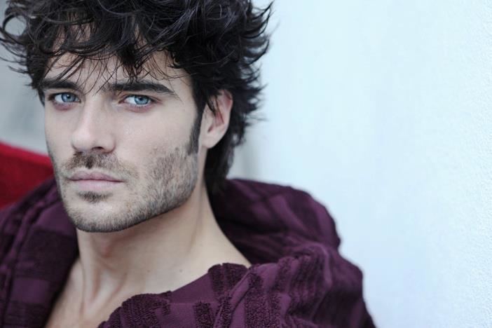 Giulio Berruti with a serious face, messy hair, blue eyes, mustache, and beard while wearing a violet robe