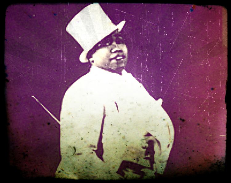 Gladys Bentley is serious, standing, has bald hair, wearing a white top hat, black earing, and a white coat.