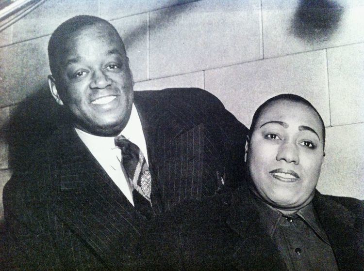 On the left, Bob Howard is smiling, leaning to his right, has bald hair, wearing a white polo with black tie under a black coat, at the right, Gladys Bentley is smiling, sitting in front of Bob Howard, has bald hair, wearing black polo and black coat.