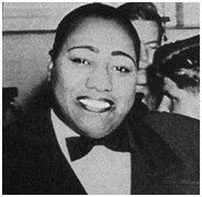 Gladys Bentley is smiling, has bald black hair, wearing a white polo with black bowtie under a black tuxedo.