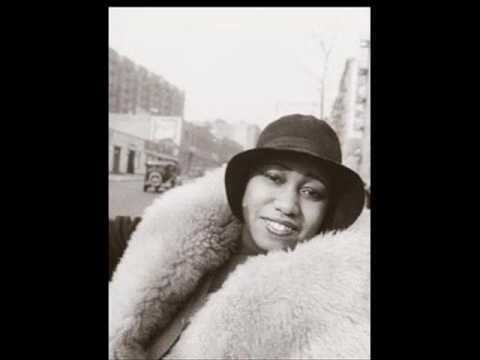 Gladys Bentley is smiling, leaning to her left, behind her is a road with a black car, wearing black cloche hat, white winter fur jacket.