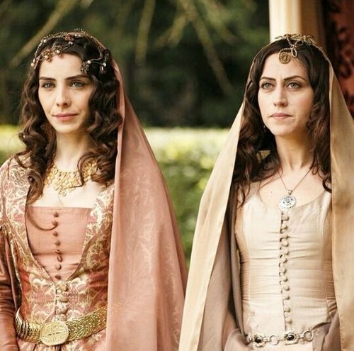 Selen Ozturk in Magnificent Century TV Series as Gülfem Hatun wearing a necklace and a brown dress with a woman wearing a necklace and a pink dress.