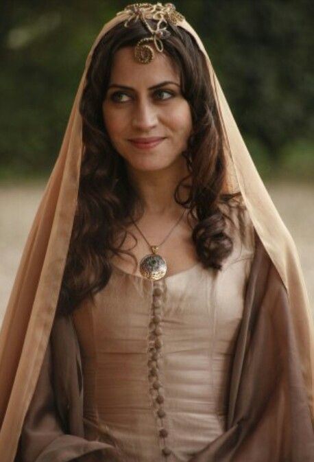 Selen Ozturk with long curly hair in Magnificent Century TV Series as Gülfem Hatun wearing a necklace and a brown dress.