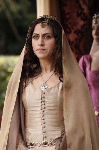 Selen Ozturk in Magnificent Century TV Series as Gülfem Hatun wearing a necklace and a brown dress with a girl on her back wearing a purple dress.