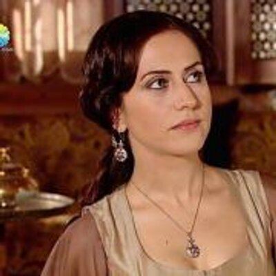 Selen Ozturk in Magnificent Century TV Series as Gülfem Hatun wearing earrings, a necklace, and a brown dress.