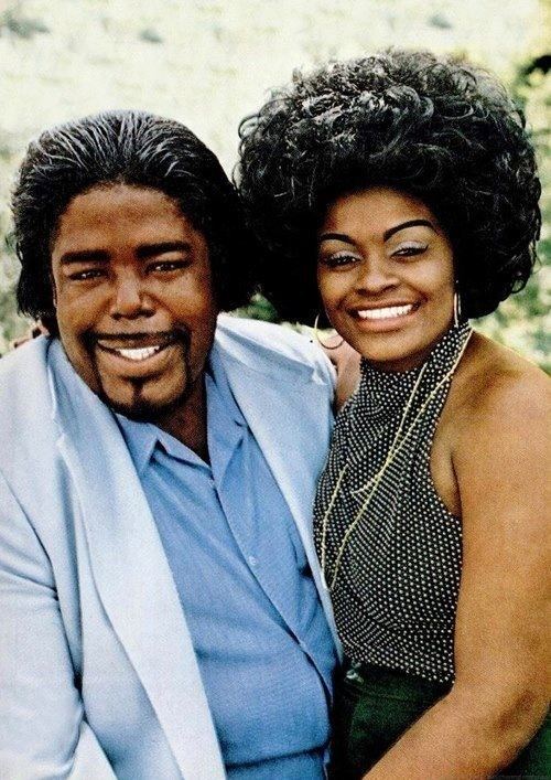 Barry and Glodean White are smiling. Barry with beard and mustache, wearing a light blue coat over blue long sleeves while Glodean with kinky hair, wearing hoop earrings, necklace, a black halter top, and black skirt.