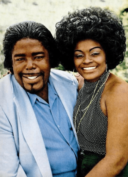Barry and Glodean White are smiling. Barry with beard and mustache, wearing a light blue coat over blue long sleeves while Glodean with kinky hair, wearing hoop earrings, necklace, a black halter top, and black skirt.