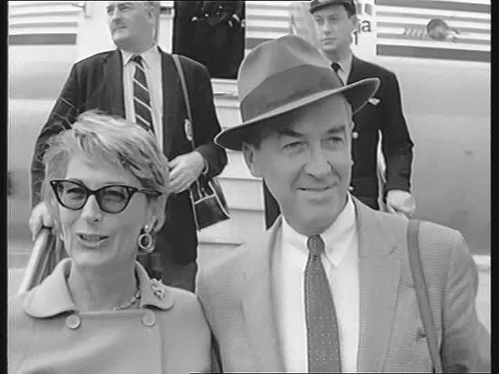 Gloria Hatrick McLean smiling while walking down the stairs of an airplane with James Stewart wearing a hat, coat, long sleeves, and necktie